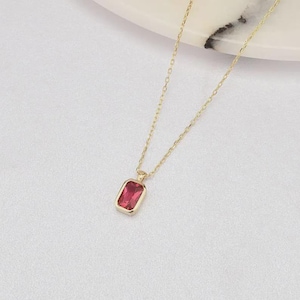 May include: A gold-colored necklace with a rectangular, ruby-red gemstone pendant. The pendant is set in a gold bezel and hangs from a delicate gold chain. The necklace is displayed on a white surface.