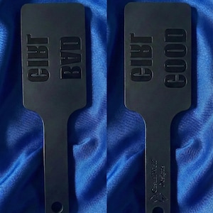 Puede incluir: Dos paletas negras con texto. Una paleta dice "GIRL BVD", la otra "GIRL GOOD". Las paletas tienen un mango con un agujero en el extremo. El texto "Casual Wolf Designs" está impreso en el mango.