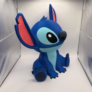 May include: A blue figurine of the character Stitch from Lilo & Stitch. The figurine has large ears with pink inner ears, a light blue belly, and a dark blue nose and claws. The figurine is sitting down.
