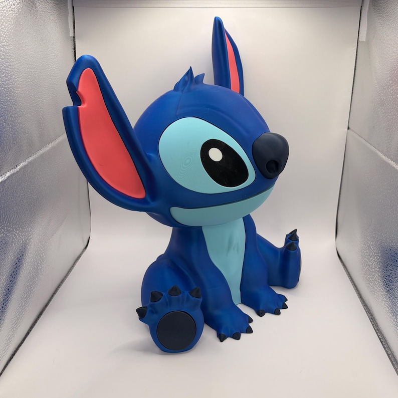 May include: A blue figurine of the character Stitch from Lilo & Stitch. The figurine has large ears with pink inner ears, a light blue belly, and a dark blue nose and claws. The figurine is sitting down.