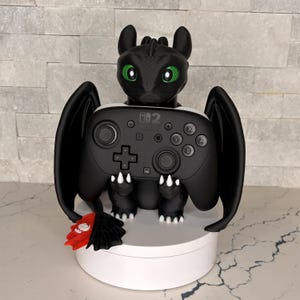 May include: A black dragon figurine with green eyes holding a black Nintendo Switch Pro controller. The dragon has large wings and a red and black tail fin. The controller features the Nintendo Switch logo and standard buttons, joysticks, and a D-pad.