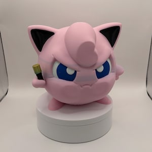 May include: A pink, round figurine with blue eyes and black-tipped ears, holding a small, gold and black object. The figurine is on a white, circular base. The overall design is reminiscent of a popular cartoon character.