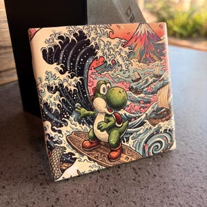 May include: A square, decorative item featuring a cartoon Yoshi character surfing a wave. The design incorporates elements of Japanese art, with a mountain, boats, and stylized waves in shades of blue, pink, and orange.