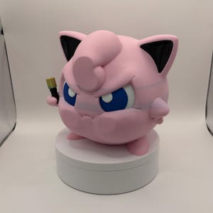May include: A pink, round figurine with blue eyes and black-tipped ears, holding a small, gold-colored object. The figurine is on a white, circular base. The overall design is reminiscent of a popular cartoon character.