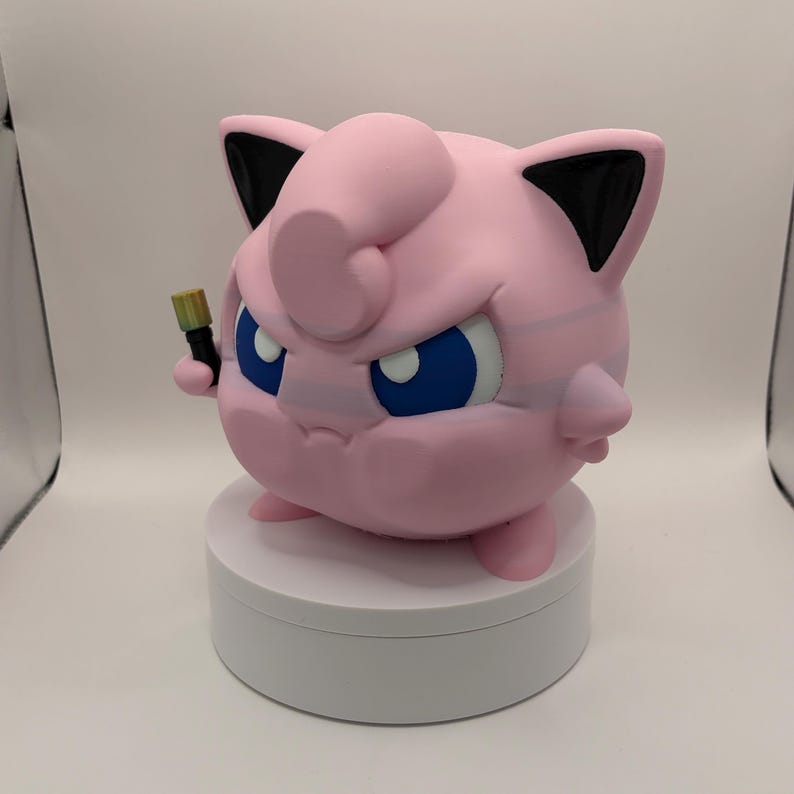 May include: A pink, round figurine with blue eyes and black-tipped ears, holding a small, gold-colored object. The figurine is on a white, circular base. The overall design is reminiscent of a popular cartoon character.