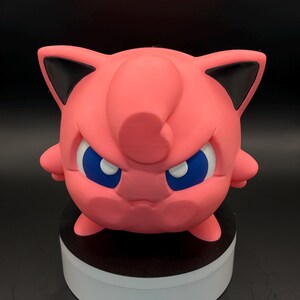May include: A pink, 3D-printed figure of a cartoon character with a round body, small arms, and pointed ears with black tips. The character has blue eyes and a stern expression. The figure is displayed on a white and black circular base.