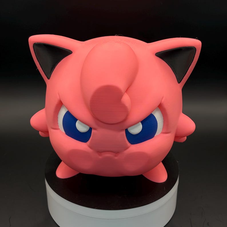 May include: A pink, 3D-printed figure of a cartoon character with a round body, small arms, and pointed ears with black tips. The character has blue eyes and a stern expression. The figure is displayed on a white and black circular base.