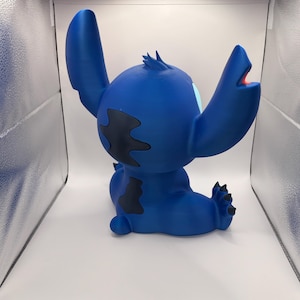 May include: A blue figurine of a cartoon character with large ears, black markings, and a wide open mouth. The figurine is sitting down with its arms raised. The character's eyes are light blue and its mouth is red.