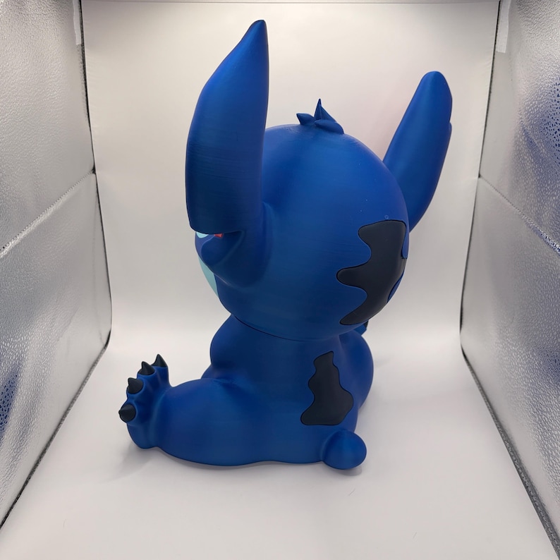 May include: A blue figurine of the character Stitch from Lilo & Stitch. The figurine is in a seated position with large ears, dark blue patches, and a small tuft of hair. The figurine is a solid blue color with black accents.