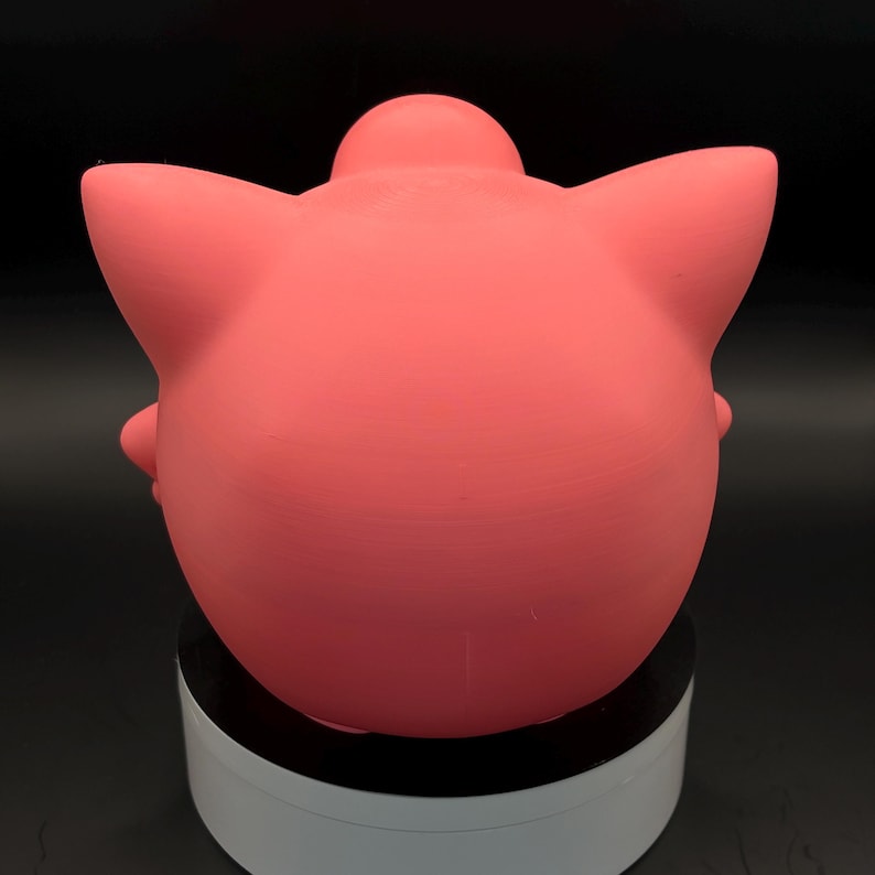 May include: A solid pink, rounded figurine with pointed ears and a small, rounded protrusion on top. The figurine is smooth and appears to be made of a solid material. The object is sitting on a white and black rotating stand.