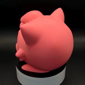May include: A pink, rounded figurine with pointed ears and a small, protruding feature on its side. The figurine is a solid color and appears to be made of a smooth material. The object is sitting on a white and black circular base.