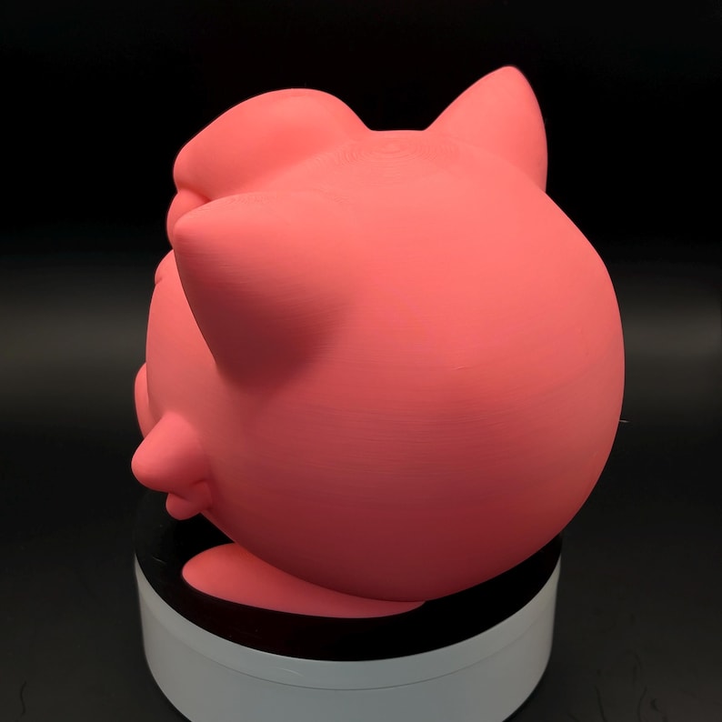 May include: A pink, rounded figurine with pointed ears and a small, protruding feature on its side. The figurine is a solid color and appears to be made of a smooth material. The object is sitting on a white and black circular base.