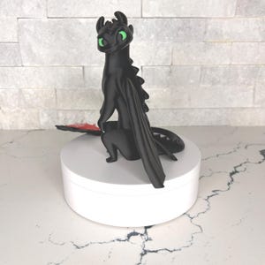 Op de afbeelding: Een zwart drakenfiguur met felgroene ogen, puntige oren en een gladde, matte afwerking. Het wezen is afgebeeld in een zittende houding met gevouwen vleugels en een lange staart, staand op een verhoogde, effen witte ronde basis.