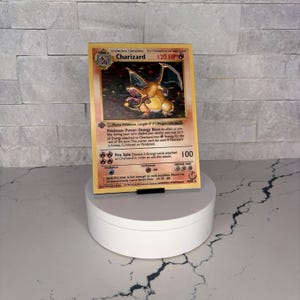 May include: A Charizard Pokémon trading card. The card features an orange dragon with black wings. The card has text that reads "Charizard 120 HP" and other game information. The card is displayed on a white rotating stand.