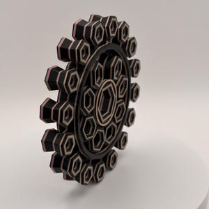 May include: A layered, geometric wooden art piece. The design features a circular shape with repeating hexagonal patterns in black, light brown, and pink. The piece is three-dimensional, with each layer adding depth and dimension to the design.