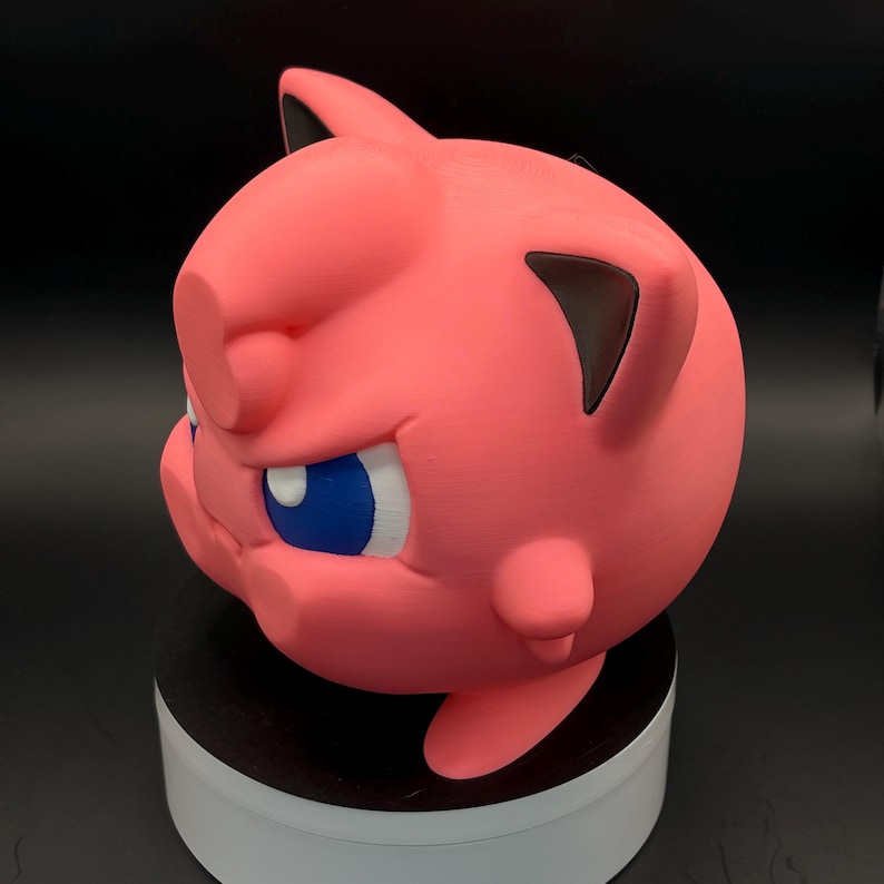 May include: A pink, 3D-printed figure of a cartoon character with blue eyes and black ears. The figure is round with a slightly angry expression. The figure is sitting on a white and black circular base.