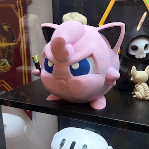 May include: A pink, 3D-printed Jigglypuff figure with blue eyes and black-tipped ears, holding a small gold object. The figure is on a black shelf, with other decorative items in the background.