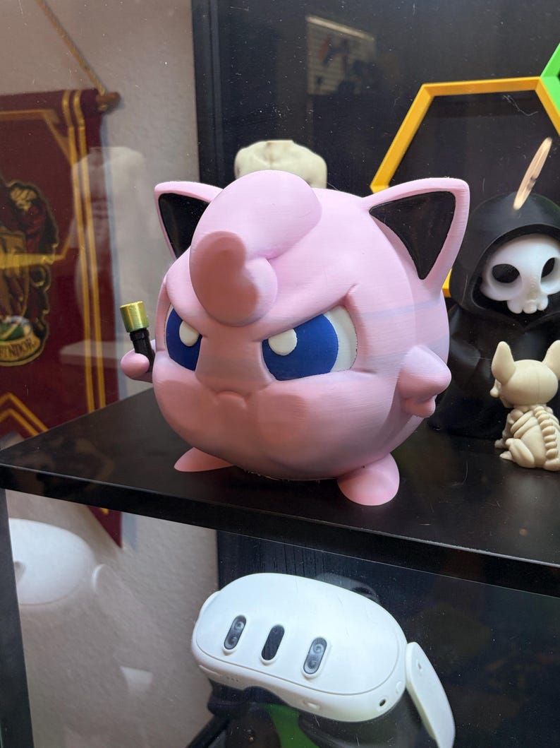 May include: A pink, 3D-printed Jigglypuff figure with blue eyes and black-tipped ears, holding a small gold object. The figure is on a black shelf, with other decorative items in the background.