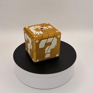 May include: A gold-colored puzzle cube with white question marks on each side. The cube is sitting on a black and white rotating platform. The cube is a replica of the iconic question mark block from the Super Mario video game series.