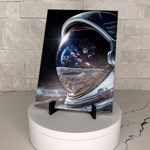 Puede incluir: Una impresión rectangular que muestra un casco de astronauta detallado con una visera reflectante. La visera muestra una escena cósmica con una vista de la superficie lunar, un planeta azul distante, una galaxia brillante y estrellas.