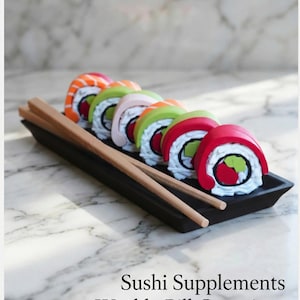 May include: A black rectangular pill container shaped like sushi rolls, with wooden chopsticks. The sushi rolls are colorful, with red, orange, green, and pink accents. The text "Sushi Supplements Weekly Pill Container" is at the bottom.