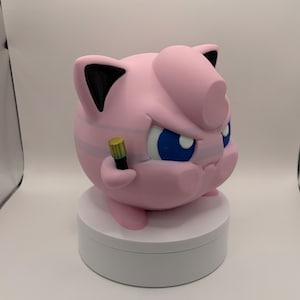 May include: A pink, 3D-printed figure of a cartoon character with blue eyes, black ears, and a determined expression. The figure is holding a small, gold-colored microphone and is posed on a white, circular platform.