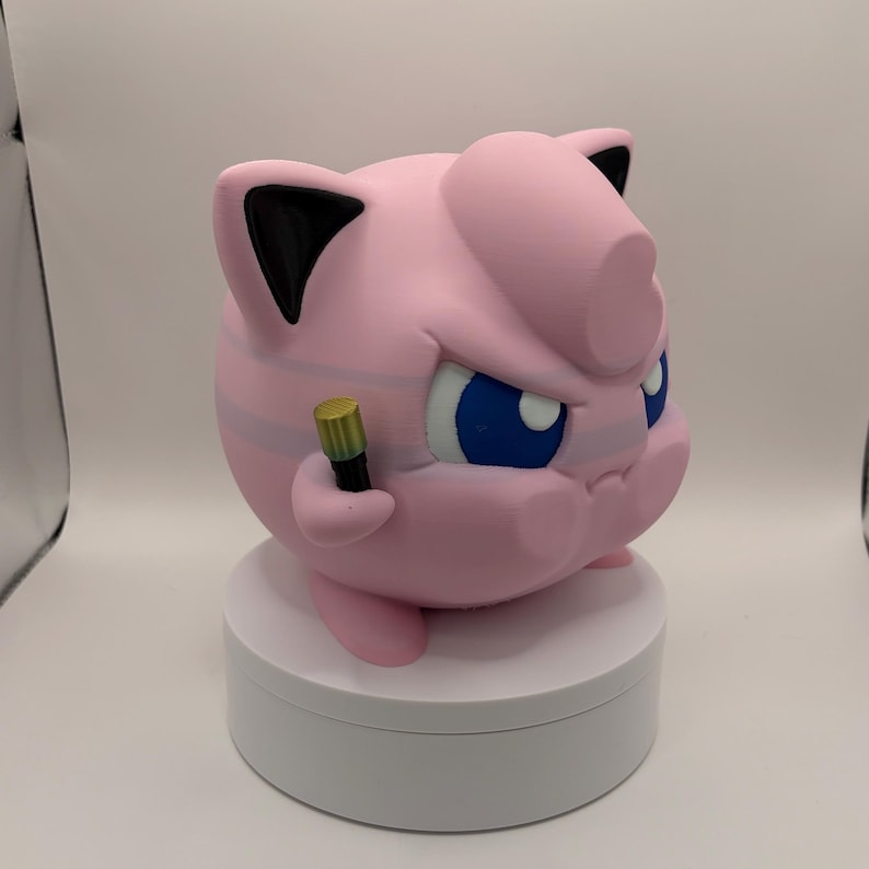 May include: A pink, 3D-printed figure of a cartoon character with blue eyes, black ears, and a determined expression. The figure is holding a small, gold-colored microphone and is posed on a white, circular platform.