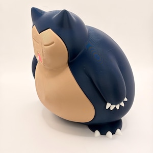 Könnte beinhalten: Eine marineblaue und beige Figur von einem schlafenden Snorlax. Die Figur hat geschlossene Augen, einen rosa Mund und weiße Krallen. Die Figur ist ein beliebter Charakter aus dem Pokémon-Franchise.