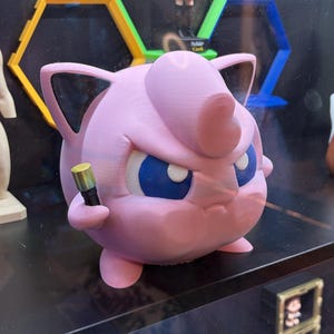 May include: A pink 3D-printed figure of a Jigglypuff-like character, with blue eyes and a stern expression, holding a small, gold-colored object. The figure is displayed on a shelf with other items in the background.