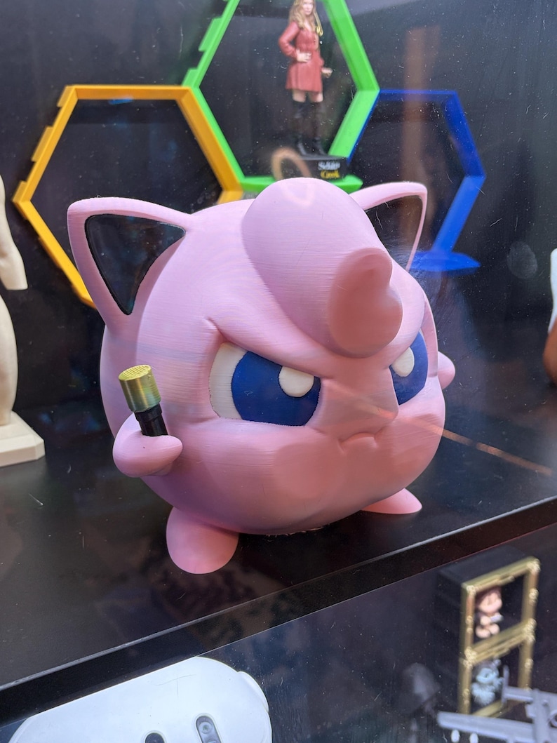May include: A pink 3D-printed figure of a Jigglypuff-like character, with blue eyes and a stern expression, holding a small, gold-colored object. The figure is displayed on a shelf with other items in the background.