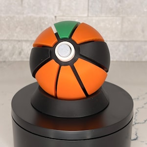 Op de afbeelding: Een bolvormig puzzelspeelgoed met gesegmenteerde panelen in oranje, zwart en groen. Het object heeft een centrale witte ronde knop aan de bovenzijde en is gemonteerd op een zwarte ronde voet, wat een veelkleurig geometrisch ontwerp laat zien.