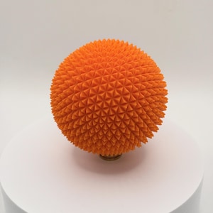Könnte beinhalten: Eine orangefarbene Kugel mit einer strukturierten Oberfläche aus dreieckigen Spitzen. Das dekorative Objekt steht auf einem kleinen, runden, goldfarbenen Sockel. Die Kugel hat einen leuchtenden Orangeton.
