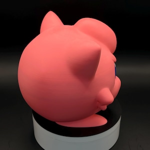 May include: A pink, three-dimensional figure of a cartoon character with large eyes and pointed ears. The figure is set against a dark background and appears to be made of a solid material. The character's face is visible, with blue eyes and a small mouth.