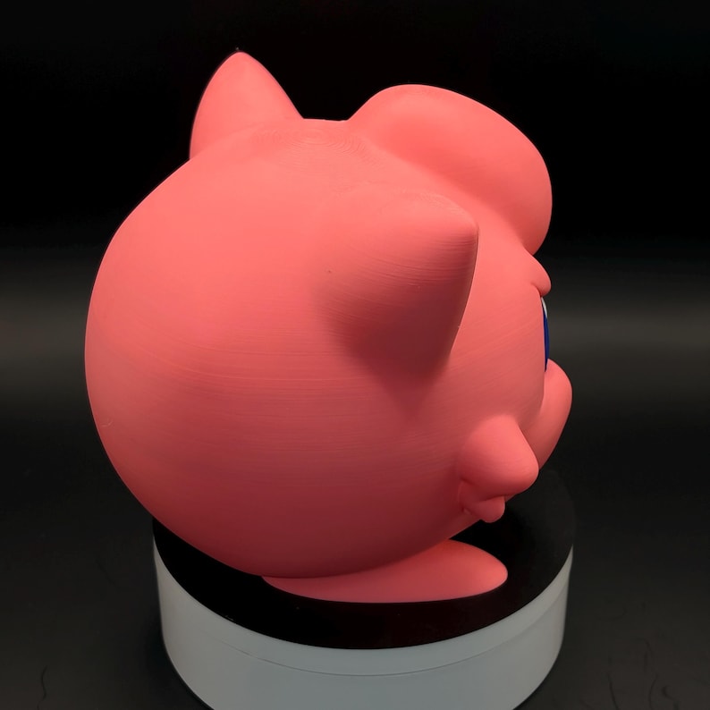 May include: A pink, three-dimensional figure of a cartoon character with large eyes and pointed ears. The figure is set against a dark background and appears to be made of a solid material. The character's face is visible, with blue eyes and a small mouth.