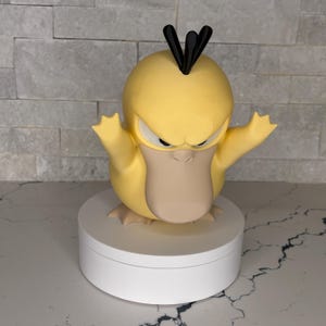Könnte beinhalten: Eine gelbe, entenähnliche Charakterfigur mit einem beigefarbenen Schnabel, drei schwarzen Haarbüscheln auf dem Kopf und einem wütenden Gesichtsausdruck. Die Figur hat die Arme erhoben und steht auf einem glatten, weißen, zylindrischen Sockel.