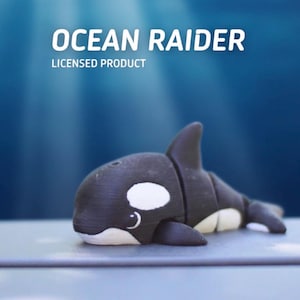 May include: A black and white orca figurine with the text "OCEAN RAIDER" and "LICENSED PRODUCT" in white. The orca has a white patch near its eye and belly. The background is a gradient of blue.