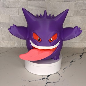 Könnte beinhalten: Eine violette Gengar-Figure mit spitzen Ohren, roten Augen, einem breiten weißen Grinsen und einer langen rosa Zunge, die aus dem Mund ragt. Die Figur ist auf einem runden weißen Sockel montiert und weist eine glatte, matte Oberfläche auf.