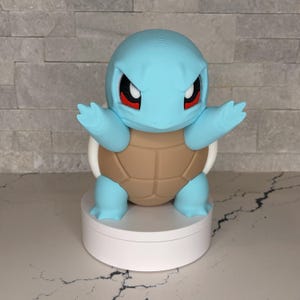 Könnte beinhalten: Hellblaue Squirtle-Figur mit braunem Panzer und weißen Akzenten. Die Figur hat rote Augen und ist mit ausgestreckten Armen posiert. Sie wird auf einem weißen, runden Sockel präsentiert.