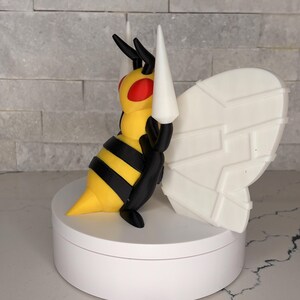 Könnte beinhalten: Eine dreidimensionale Bienenfigur mit einem gelb-schwarz gestreiften Körper, roten Augen und schwarzen Fühlern. Das Insekt hat zwei große, weiße, geometrische Flügel mit paneelartigen Details und steht auf einem runden weißen Sockel.