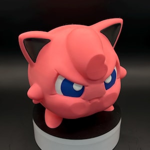May include: A pink 3D-printed figure of a cartoon character with blue eyes and black-tipped ears. The figure has a round body and a slightly angry expression. The figure is sitting on a white and black base.
