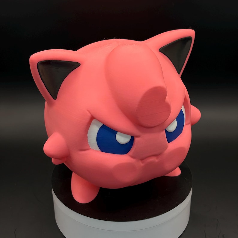 May include: A pink 3D-printed figure of a cartoon character with blue eyes and black-tipped ears. The figure has a round body and a slightly angry expression. The figure is sitting on a white and black base.