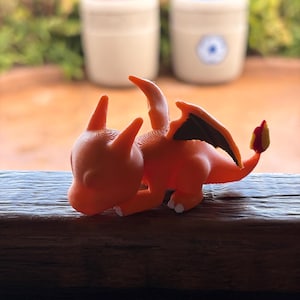 May include: A 3D-printed orange Charizard figurine, a popular character from the Pokémon franchise. The figurine is lying down, with its wings and tail visible. The tail has a yellow and red flame. The figurine is on a wooden surface.