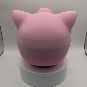 May include: A pink, spherical figurine with large ears and a small, rounded top. The figurine has two light purple stripes around its body. It is displayed on a white, circular stand against a white background.