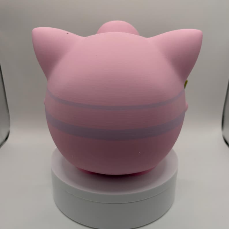 May include: A pink, spherical figurine with large ears and a small, rounded top. The figurine has two light purple stripes around its body. It is displayed on a white, circular stand against a white background.