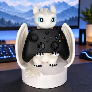May include: A white dragon figurine with large blue eyes and spread wings, designed as a controller stand. The dragon holds a black Xbox wireless controller with a directional pad, two analog sticks, and colored action buttons in its claws.