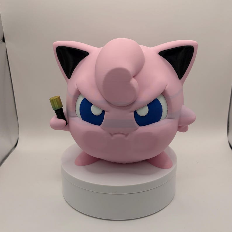 May include: A pink, round figurine with blue eyes and black-tipped ears, holding a small, black and gold object. The figurine is on a white, circular base. The character has an angry expression.
