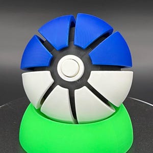May include: A 3D-printed spherical puzzle with blue and white segments, set on a green base. The puzzle's design features a series of curved, interlocking pieces. The central core is black with a white circular element.