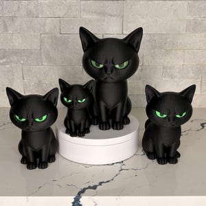 Op de afbeelding: Een set van vier zwarte kattenbeeldjes met getextureerde oppervlakken en felgroene ogen. Eén grote kat zit op een witte ronde sokkel, terwijl drie kleinere katten eromheen zijn geplaatst, allemaal met identieke norse gezichtsuitdrukkingen.