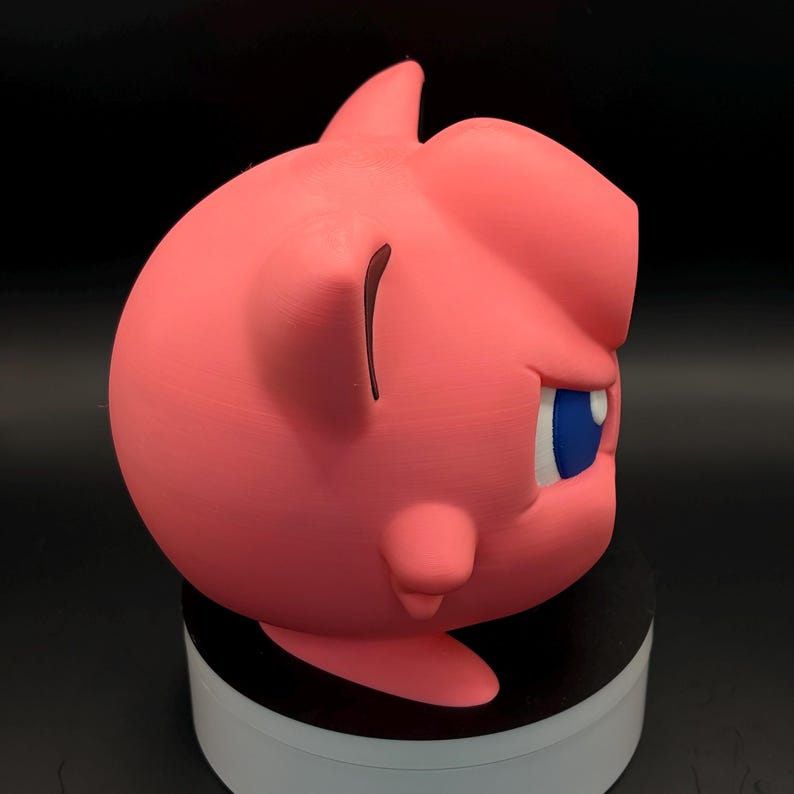 May include: A pink, 3D-printed sculpture of a cartoon character's head, featuring a prominent fin-like structure on top. The character has a blue eye, a small ear, and a rounded shape. The sculpture is displayed on a black surface.