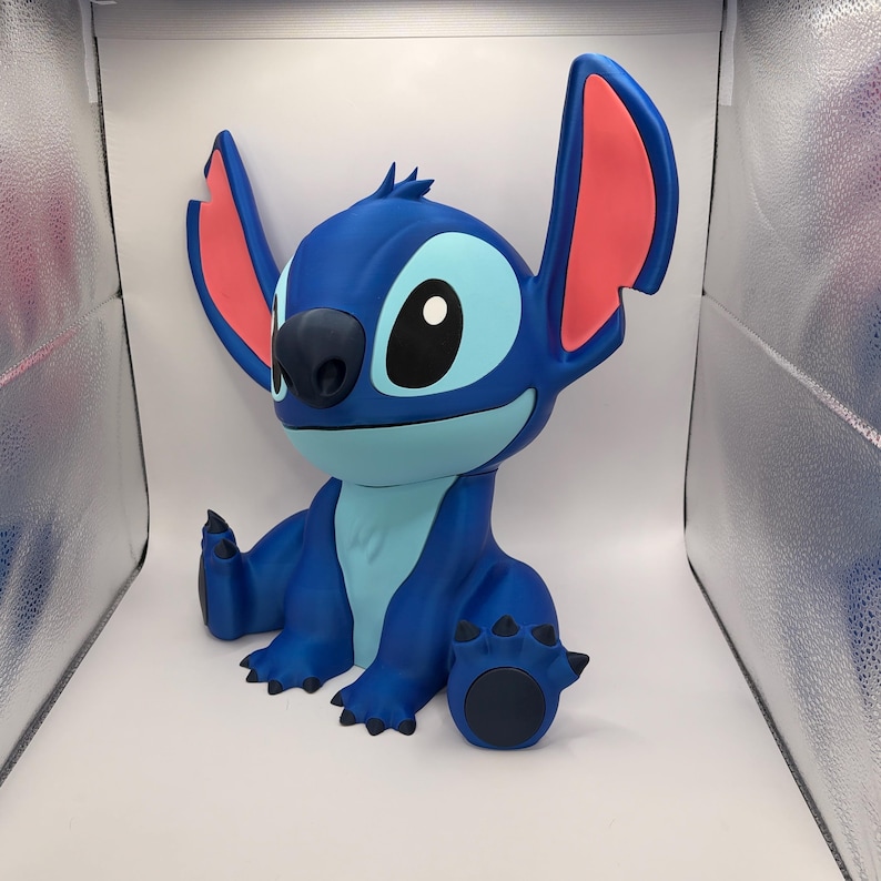 May include: A blue Stitch figurine with large ears, a black nose, and large eyes. The figurine is sitting with its arms and legs outstretched. The inside of the ears are pink. The figurine is a popular character from the animated movie Lilo & Stitch.